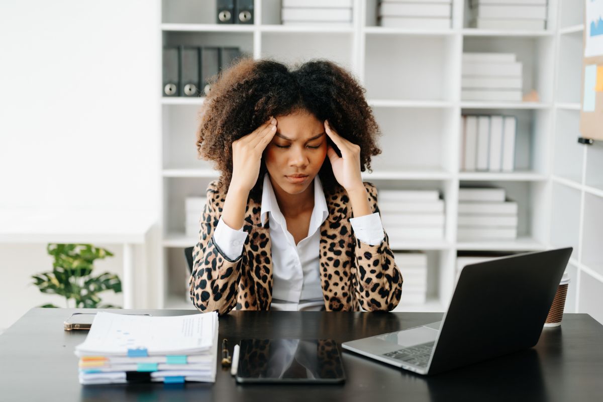 A professional Black woman sitting at a desk, looking exhausted behind the scenes, illustrating the high cost of ADHD masking at work.